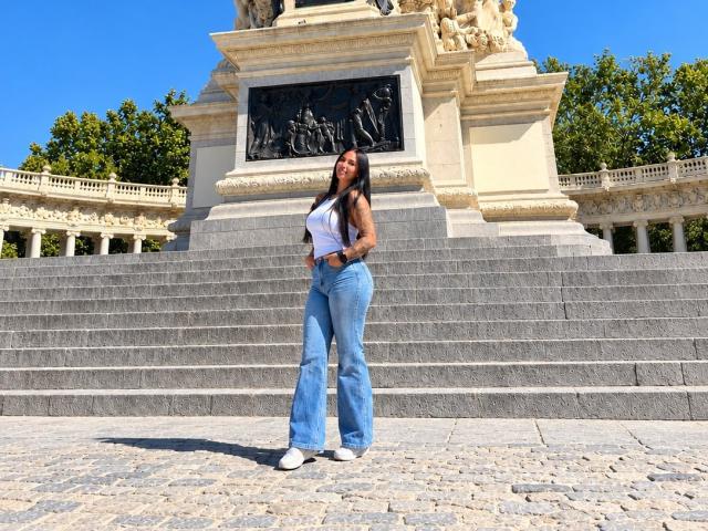 AnastasiaVolkov standing in front of a tall monument, wearing a white tank top, blue flared jeans, and white sneakers, with long black hair and a smiling face.