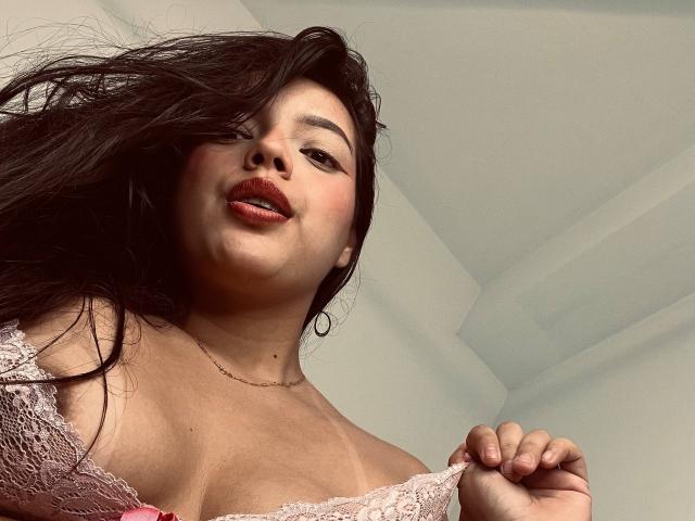 ViolettaRouse with long black hair, red lipstick, and lace pink bra, pulling strap down, showing big boobs, looking down at camera, light skin, hoop earrings, subtle smile.