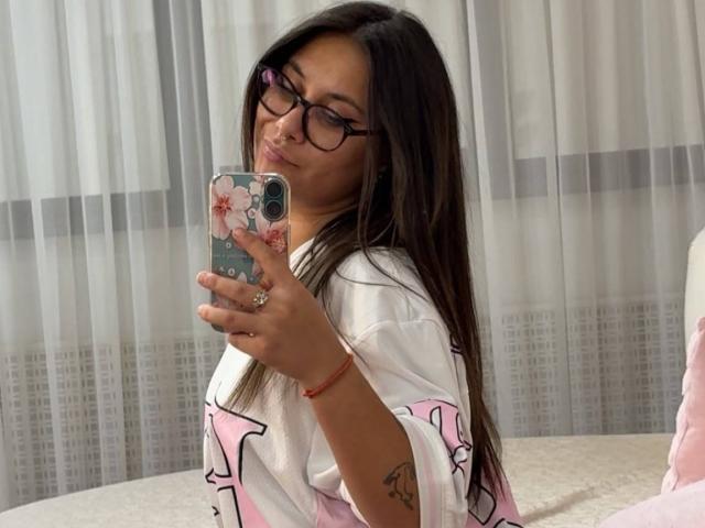 JessieBoo con lunghi capelli castani, occhiali, camicia bianca con motivi rosa e neri, che tiene in mano un telefono con una custodia a fiori, che bacia il suo riflesso, con un piccolo tatuaggio sul braccio destro, seduta sul letto..