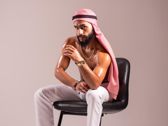 TahielFoster, sem camisa, com óleo brilhante nos músculos, keffiyeh xadrez vermelho, cadeira preta, calças brancas, colar de ouro, relógio de pulso, barba, expressão pensativa, sentado, pele morena, sexy, atraente..