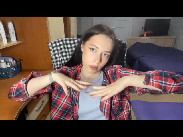 Evangeline Haught, avec sa peau pâle, ses yeux bleus et ses cheveux brun foncé, portant une chemise à carreaux rouges par-dessus un haut bleu, les mains sur la poitrine, avec une expression légèrement agacée, dans une chambre..
