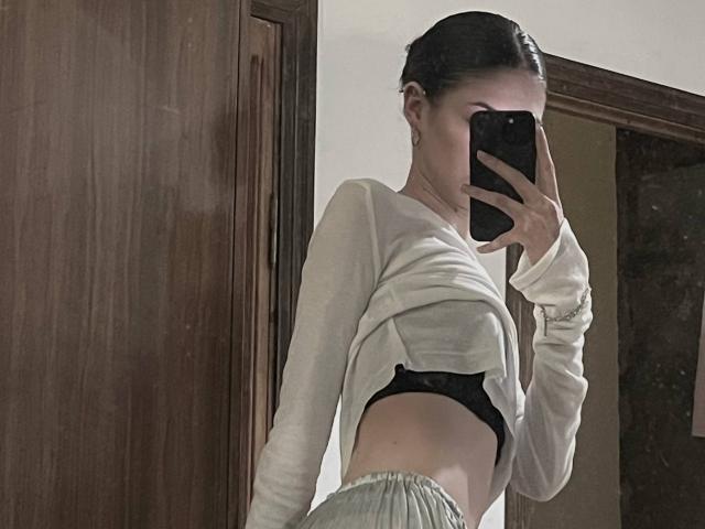 MonaMarchetta with dark hair in a braid, wearing a white long-sleeve shirt and gray pants, showing black bra, taking a mirror selfie with her phone.
