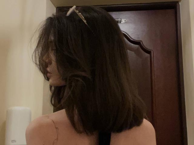 MonaMarchetta with messy dark brown hair, wearing a black strap, facing sideways, showing her bare back and a tattoo on her left shoulder, in a dimly lit room.