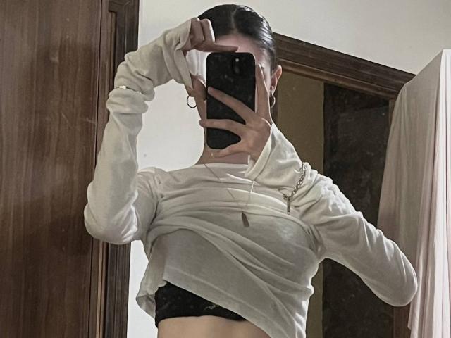 MonaMarchetta lifting a white hoodie to reveal black lace underwear, covering her face with a phone, her hand in her dark hair. Her tits are partially visible.