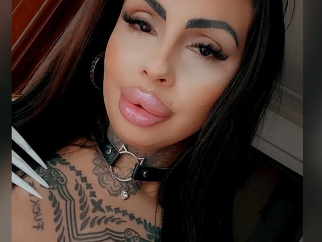 DenyChantal with glossy pink lips, black eyebrows, long eyelashes, black hair, black leather collar, tattooed chest, and hoop earrings, looking at the camera with a slight pout.
