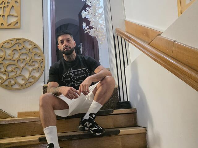 TommyDum, con barba, collo tatuato, cappello nero, maglietta nera, pantaloncini bianchi, calze bianche, scarpe da ginnastica nere e bianche, seduto su delle scale di legno, con un'espressione leggermente infastidita..