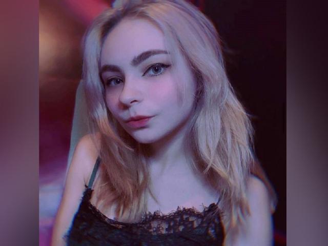 CabriaElles, con cabello rubio, ojos azules y un rostro adorable, vestida con una blusa negra de encaje, tiene un leve ceño fruncido y una expresión ligeramente melancólica en una fotografía con una iluminación suave..