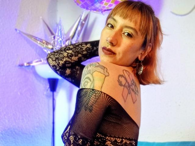 AliceRiot met kort bruin haar, een neusring, donkere lipstick, tatoeages op haar schouder, gekleed in een zwarte kanten top en een top van netstof, kijkt over haar schouder, staand in een kamer met een paarse lamp..
