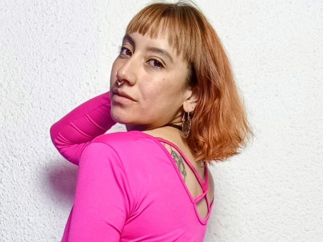 AliceRiot with short, orange-dyed hair, pink nose ring, and pink top, showing tattoos on her neck, wearing large earrings, looking over her shoulder. White textured wall.