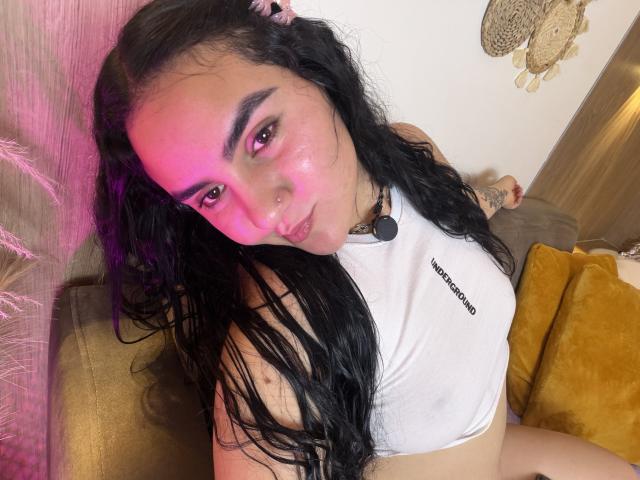 FenixAlthea with long black hair, pink blush, and white top, looking at camera, nose piercing, pink light, yellow pillow, brown arm, and tattoo on right arm.
