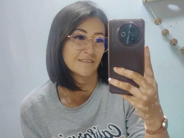 SharissWilliam with a bob haircut, glasses, nose ring, and grey t-shirt, taking a mirror selfie with a pink phone, showing a subtle smile and painted nails.