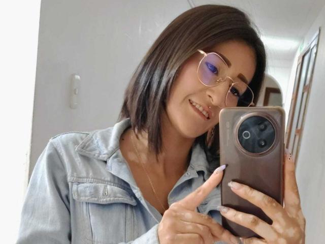 SharissWilliam with shoulder-length brown hair, wearing glasses and a denim shirt, holding a phone, smiling at the mirror, showing her ass with a gold necklace.