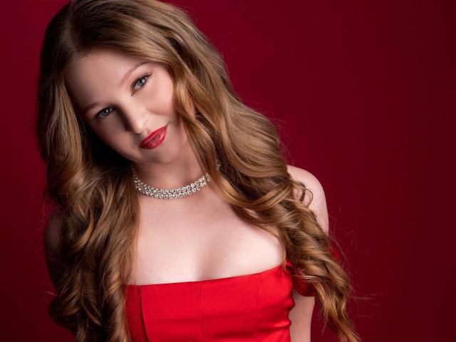 AkilahGhil with long, wavy blonde hair, red dress, and sparkling necklace, smiling at the camera, showing her tits against a dark red background.