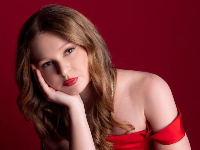 AkilahGhil with wavy blonde hair, red lipstick, and off-shoulder red dress, resting her chin on her hand, looking seductively at the camera, against a red background.