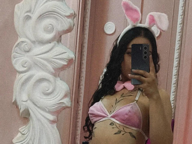 MiiaBennet in pink velvet bra with bunny ears, holding phone in mirror selfie. Long curly black hair, tan skin, small pink bow tie, visible tattoo on right side.