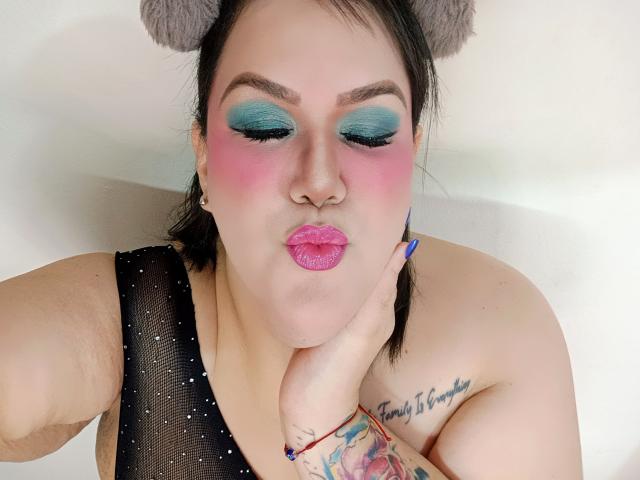 GiiannaMorrys com sombra azul-esverdeada, blush rosa e batom rosa, fazendo beicinho com uma mão no rosto. Ela tem tatuagens, uma blusa de malha preta e orelhas de urso cinzas..