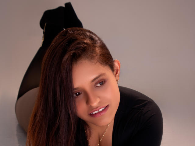 ValerieSynclair, com cabelos castanhos longos, blusa preta e botas pretas, deitada de bruços, sorrindo, mostrando seus pequenos seios e nádegas, contra um fundo simples..