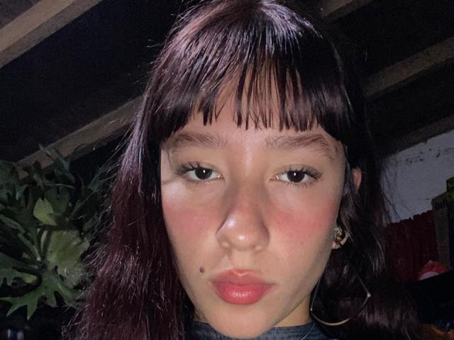 JuliethCourtouis has dark brown bangs, straight black hair, light brown skin, small nose mole, red lips, brown eyes, and small earrings. She's looking at the camera with a serious expression.