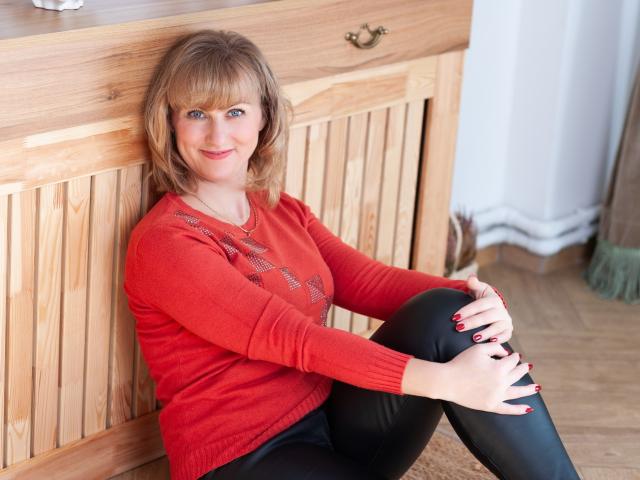 RebecaMarti with blonde hair, red sweater, black leather pants, sitting against wooden cabinet, smiling, hands on knees, red nails, wooden floor, brass handle, soft light, relaxed.