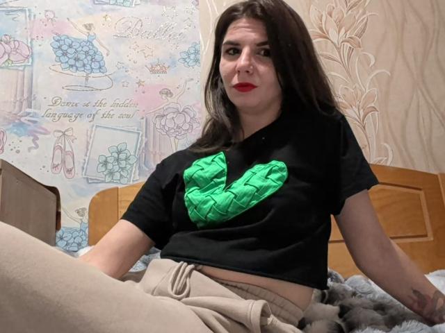 VickiLipoma with dark brown hair, red lipstick, black shirt with green handprints, beige pants, sitting on bed, looking up with slightly annoyed expression. Her ass is partially visible.
