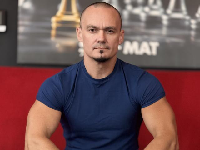 KaelSage, chauve avec une petite barbe, musclé, portant un t-shirt bleu moulant, assis les bras croisés, expression sérieuse, mur rouge en arrière-plan..
