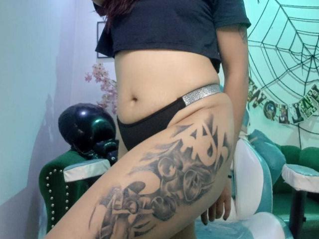 AriannaSeete's midriff showing a black crop top, black panties with a sparkly waistband, and a large tattoo on her right thigh, leaning on a green chair.
