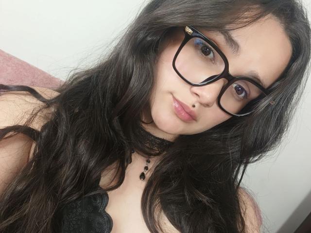 Natalia Rivas, con cabello negro largo, un collar de encaje negro, gafas negras, un piercing en la nariz y labios rosados brillantes, vestida con una blusa de encaje negro, mostrando sus senos..