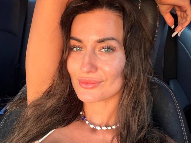 Mireille Veil heeft groene ogen, bruin golvend haar, en draagt een witte top met een kralenketting. Ze zit in een auto, met haar arm omhoog, en glimlacht met licht glanzende lippen..