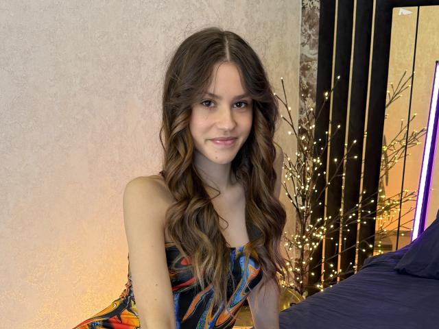 GracyMurre with long wavy brown hair, wearing a colorful strapless dress, smiling slightly with a small boogers on her nose, standing next to a bed with blue covers.