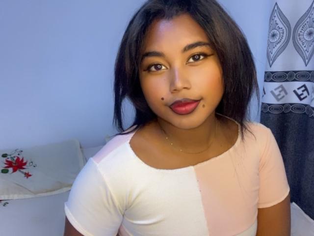 MarinahSexy with dark hair, brown eyes, and red lipstick, wearing a pink and white top, has a beauty mark on her right cheek, looking at the camera.