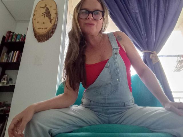 Pamela Colin, con gafas, una blusa roja y un overol vaquero azul, sentada con las piernas separadas en un sofá verde, mostrando sus pechos y un pequeño tatuaje en su brazo derecho..