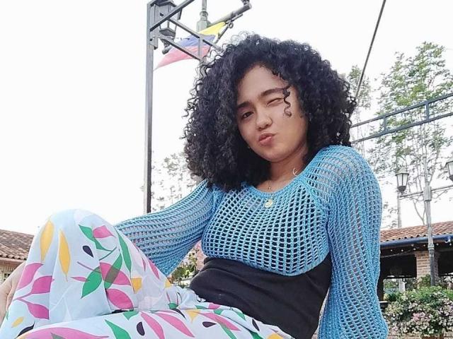 KirasPell with curly black hair, blue mesh top, and colorful floral pants, sitting with a pouty face, black waistband, and a relaxed, slightly annoyed expression.
