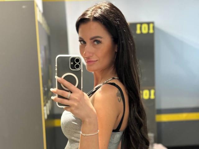 MireilleVeil with long black hair, wearing a gray tank top, showing her tits in a mirror selfie, holding a phone with a ring on her finger, smiling.