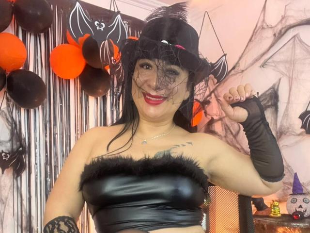 CamilaFroes with medium brown skin, black hair, small breasts, and a black lace bra and thong, standing in a bedroom, smiling, touching her hair.