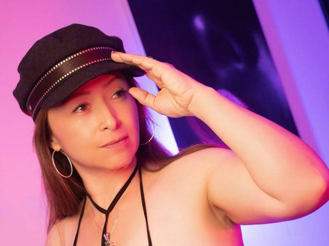 GianineLarose with a black cap, hoop earrings, and a black halter top, saluting with her right hand, has fair skin, brown hair, and a subtle smile.