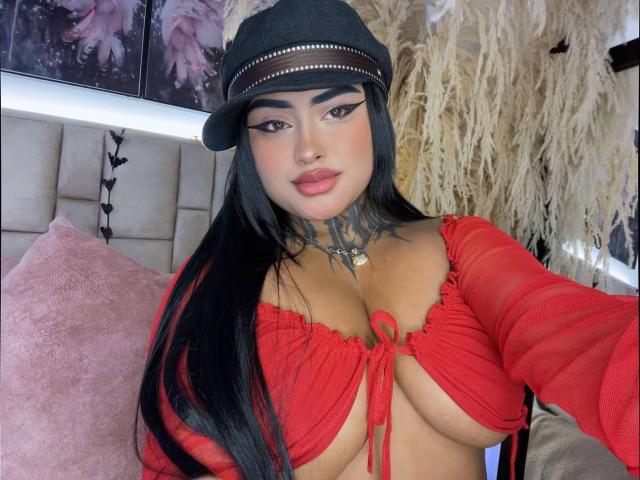 LairaHearvy with long black hair, wearing a black hat, red crop top, and white panties, showing her large tits and tattooed neck, sitting on a bed.