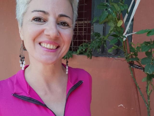 FuegoMujer tem cabelo curto e grisalho, olhos castanhos e uma camisa rosa com zíper, sorrindo com batom rosa. Ela está usando brincos longos e tem seios pequenos. Sua pele é clara..