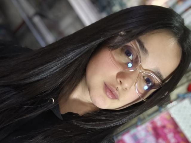 Natalia Rivas, com cabelos longos e pretos, usando óculos transparentes com luzes azuis, uma camisa preta e pele clara, olhando para a câmera com uma expressão neutra..