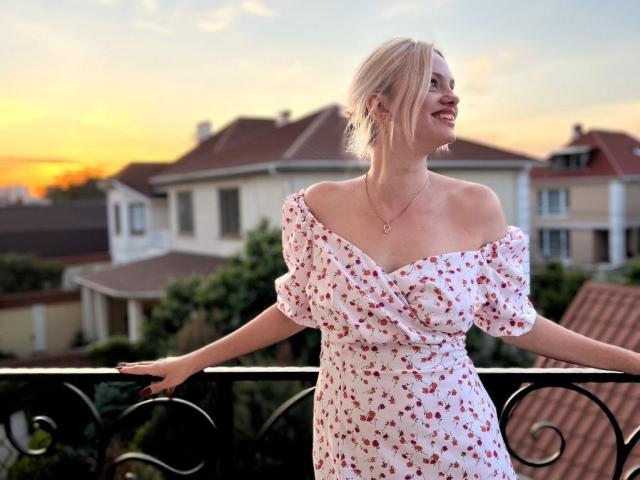 CrystalHope mit blonden Haaren, weißem Kleid mit abgesetzter Schulter und roten Blumenmustern, kleinen Brüsten, lächelnd, stehend auf einem Balkon, Sonnenuntergang im Hintergrund, haltend sich an der Reling, Halskette mit Herzanhänger..