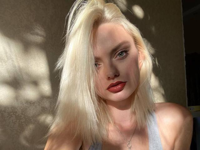 CrystalHope hat blondes Haar, helle Haut, roten Lippenstift und eine silberne Halskette. Sie trägt ein graues Tanktop, und das Sonnenlicht wirft Schatten auf ihr Gesicht..