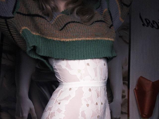 TiffaniMacandog en robe blanche en dentelle avec des motifs floraux, portant un pull rayé vert et marron, le pull relevé pour révéler ses seins, cheveux blonds, debout, robe ajustée à la taille..