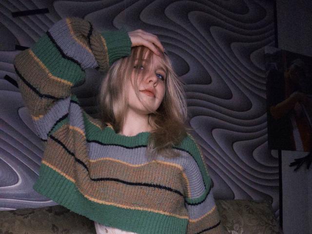 TiffaniMacandog with blonde hair, green striped sweater, hand in hair, slight smile, blue eyes, pale skin, wearing makeup, in dimly lit room, wall with wavy pattern in background.