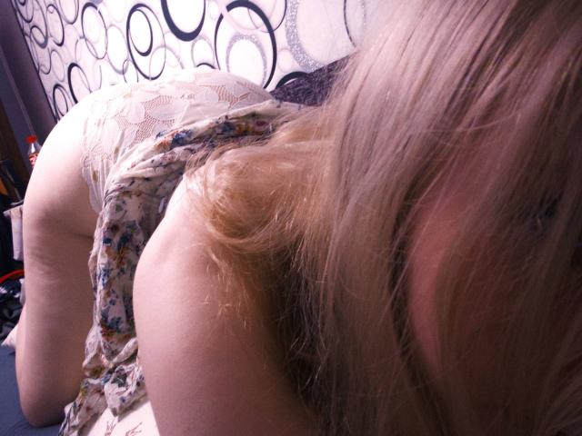 TiffaniMacandog on all fours, wearing a floral dress, her ass up, lace underwear visible, blonde hair in her face, her back and butt in focus, camera angle from above.
