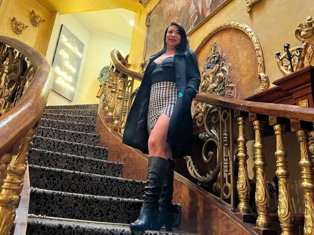Karimelee está de pie en unas escaleras ornamentadas, vistiendo un abrigo de cuero negro, una falda a cuadros y unas botas negras hasta la rodilla, mostrando su trasero, con el pelo largo y negro y una sonrisa segura..