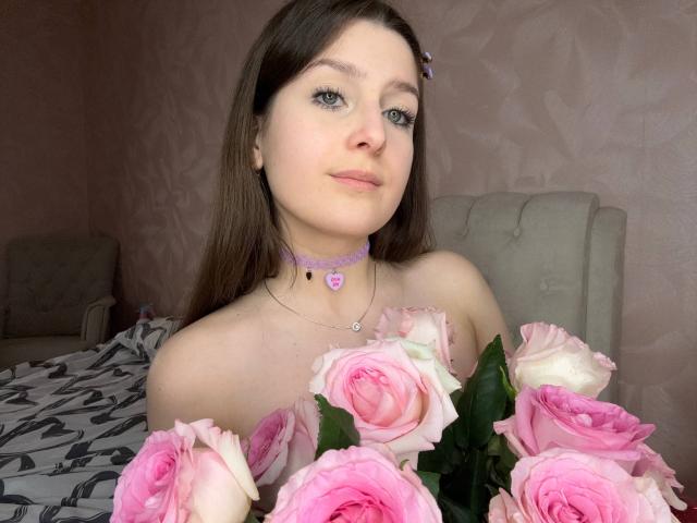 SabinaGrigalonis with pink and white roses in front of her, wearing a purple choker with a heart, in a softly lit bedroom, looking at the camera with a slight smile.
