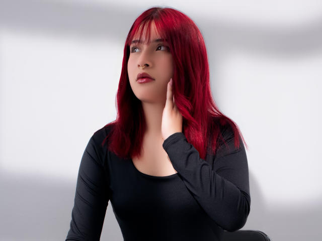 HaileyParck with red hair, wearing a black long-sleeve top, touching her face with her right hand, looking up with a slight smile, showing her tits.