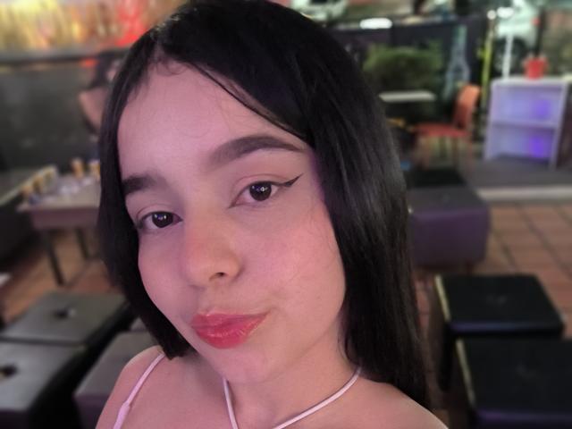 LissyLaurent, met zwart haar, roze lippenstift en een cat-eye eyeliner, frunselt haar lippen in een bar. Ze draagt een roze haltertop en heeft een schattige, vrouwelijke uitstraling..