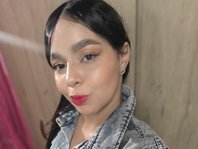LissyLaurent tem cabelo escuro, pele clara e batom vermelho, veste uma jaqueta jeans com maquiagem discreta e pequenos brincos, olhando ligeiramente para o lado..