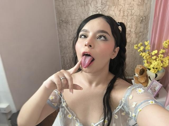 LissyLaurent with black hair in pigtails, tongue out, finger on lip, wearing sheer grey top with daisy patterns, exposing her small tits, in a cute, playful pose.