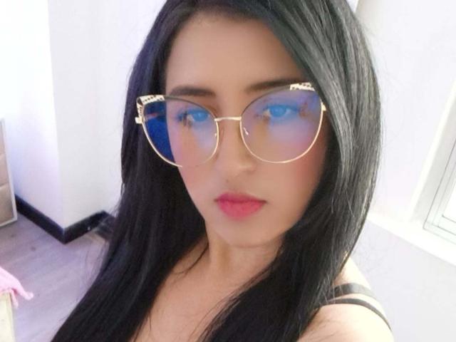 NicollJeans has long black hair, pink lips, and wears gold-rimmed glasses with blue reflective lenses, showing off her cute face and subtle makeup. Boobs are partially visible.